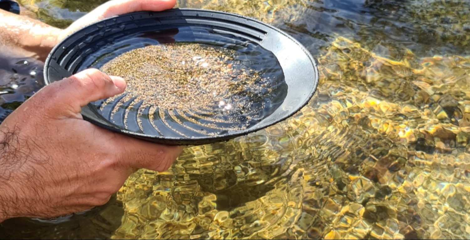 Gezeigt wird ein Ausschnit, wie eine Person an einem Fluss oder Gewässer mit einer Schale Gold wäscht